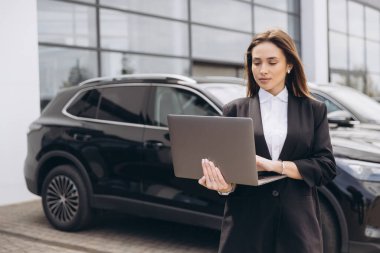 Saleswoman working on laptop in front of new cars at dealership