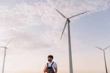 Arab engineer holding a helmet while inspecting wind turbines in a wind farm, surrounded by a breathtaking sunset landscape