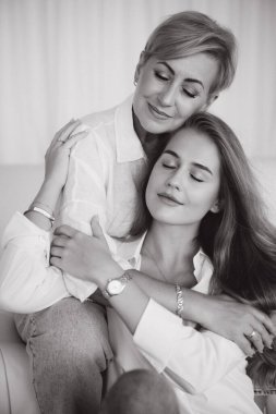 Mother and daughter are hugging each other on a sofa, both with closed eyes and smiling faces, showing a tender moment of affection and complicity