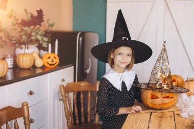 Little girl wearing witch hat and costume decorating carved pumpkins at home in kitchen for Halloween party