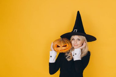 Smiling woman wearing witch hat holding carved pumpkin celebrating Halloween against yellow background