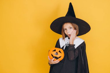 Little girl with witch costume holding a pumpkin basket with surprised expression for Halloween party on yellow background