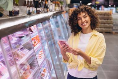 Young woman smiling while choosing meat in vacuum packaging from refrigerated shelves in supermarket