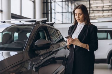 Saleswoman engaging with a smartphone while positioned next to a new vehicle in a bustling car dealership showroom