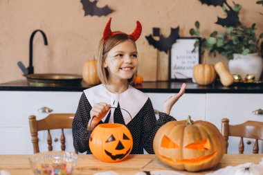 Smiling little girl wearing devil horns holding a halloween pumpkin basket and showing a jack o' lantern in a kitchen decorated for the occasion