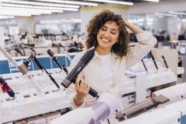 Smiling woman touching her curly hair while selecting a hairdryer among various options in an electronics store, enjoying the shopping experience