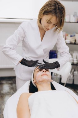 Beautician wearing black gloves applying a facial mask to a client lying on a treatment table in a professional beauty center