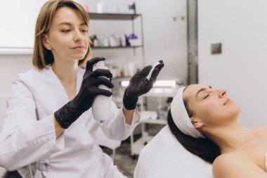Beautician wearing black gloves spraying moisturizing treatment on a client's face lying on a bed in beauty salon