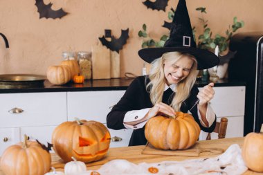 Young woman wearing a witch hat joyfully carving pumpkins for a Halloween party, surrounded by festive decorations in her cozy kitchen