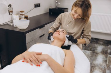 Beautician wearing black gloves cleaning face of young woman lying on massage table in beauty salon