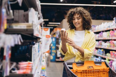 Smiling curly haired woman choosing toothpaste product in supermarket reading label information pushing shopping cart