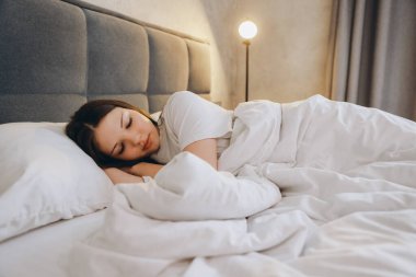 Young woman sleeping in bed with white sheets and pillows, enjoying a peaceful night sleep in a comfortable bedroom with warm light