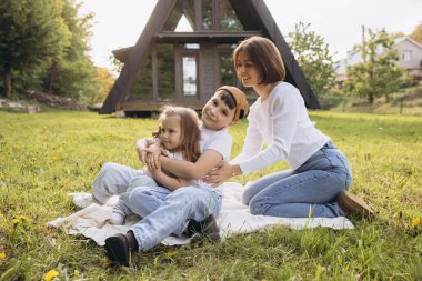 Mother and children relaxing and hugging on a blanket in the garden of their modern a frame house, enjoying quality family time