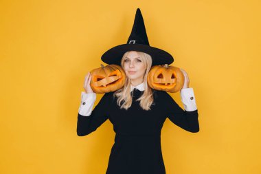 Young blonde woman wearing witch hat and black dress holding two carved pumpkins on yellow background for Halloween