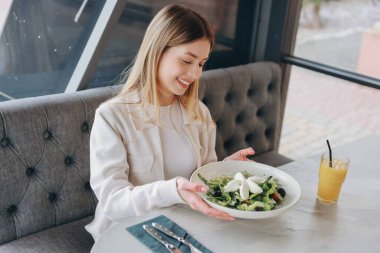 Young woman smiling and holding a plate of fresh salad in a restaurant, with a glass of orange juice on the table