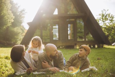 Happy family lying on lush grass, enjoying a summer vacation in front of a modern a frame cabin, creating cherished memories together