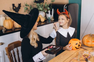 Mom with witch hat applying Halloween makeup on her daughter's face, in a kitchen decorated for the occasion