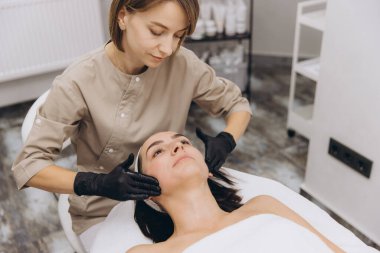 Beautician wearing black gloves massaging face of client lying on massage table in beauty salon