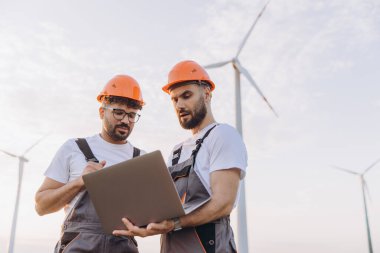 Two engineers wearing hardhats and overalls using laptop analyzing wind turbine in field for sustainable energy generation