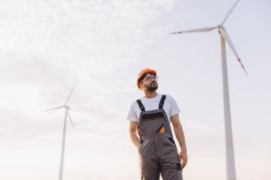 Confident arab engineer wearing overalls and hardhat inspecting wind turbines in a wind farm, ensuring sustainable energy production