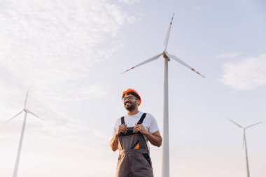 Indian engineer wearing work overalls and hardhat smiling near wind turbines generating electricity, renewable energy concept
