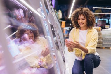 Customer selecting groceries in supermarket refrigerator aisle, holding a plastic container with food, doing grocery shopping