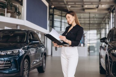 Saleswoman holding a clipboard and checking documents in a car dealership, with new cars on display