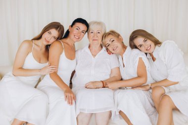 Five women of different generations wearing white dresses are sitting on a white sofa, hugging and smiling