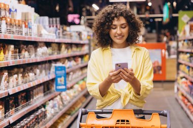 Young woman with curly hair using her smartphone while pushing a shopping cart in a supermarket