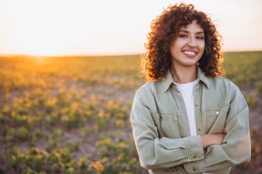 Portrait of a young agronomist smiling confidently with crossed arms, enjoying the beauty of a soybean field during a vibrant sunset