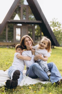 Mother hugging her two children while sitting on a blanket in the grass in front of their modern a frame house