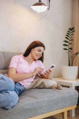 Happy woman lying on a cozy sofa in a comfortable living room, using her smartphone to browse social media and connect online