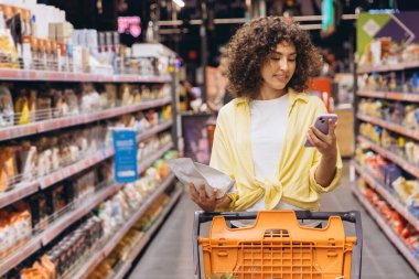 Young woman pushing shopping cart and using smartphone while choosing groceries in supermarket