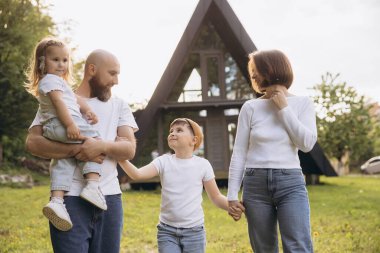 Smiling family walking and holding hands in front of their new, modern, a frame house, enjoying a sunny day together