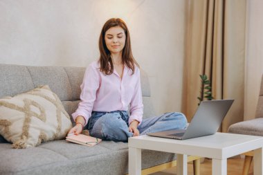 Serene young woman taking a break from work, relaxing on her comfortable sofa with a laptop and notebook in her modern apartment