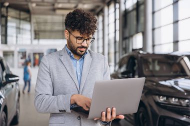 Car salesman using a laptop in a bustling car dealership, managing sales data and customer information with efficiency and focus