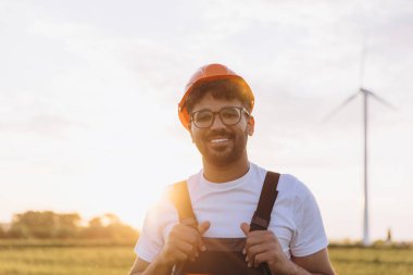Arab engineer wearing work overalls and hardhat smiling in front of wind turbine at sunset in a sustainable energy concept