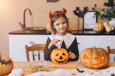 Little girl wearing devil costume is sitting at table in kitchen and holding halloween pumpkin basket