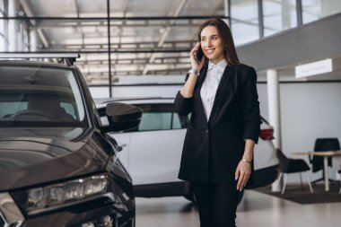 Saleswoman engaging in a phone conversation while showcasing a new car to a customer in a bustling car dealership showroom