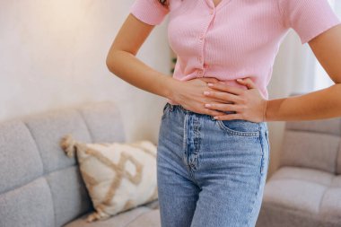 Karın ağrısı ya da karın ağrısı çeken bir kadın, elleriyle karnını tutuyor, oturma odasında duruyor.