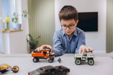 Gözlüklü akıllı çocuk evdeki oturma odasındaki masada oyuncak arabalarla oynuyor.