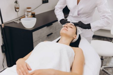 Güzellik uzmanları, güzellik salonunda genç bir kadının yüzüne krem sürerken siyah eldiven takıyor.