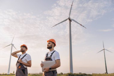 Dizüstü bilgisayar tutan iki mühendis gün batımında rüzgar türbinlerini inceliyor, sürdürülebilir enerji üzerinde çalışıyorlar.