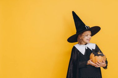 Cadılar Bayramı 'nı canlı sarı bir arka planda kutlayan bir balkabağı tutan sevimli küçük kız.