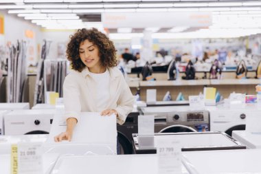 Genç bir kadın, elektronik mağazasında en iyi fiyatları ararken çeşitli çamaşır makinelerini karşılaştırıyor.