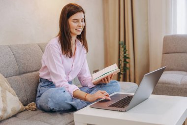 Kanepede rahat bir şekilde oturan gülümseyen kadın, kitap okurken dizüstü bilgisayar kullanıyor, uzaktan çalışıyor ve evden ders çalışıyor.