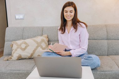 Gülümseyen kadın evde dizüstü bilgisayar kursları alırken not defterine notlar yazıyor.