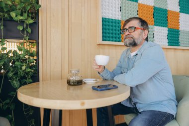 Sakallı, serbest çalışan, modern bir kafede bir fincan çayın tadını çıkaran, işe ara veren ve düşünceli bakışlarını kaçıran biri.