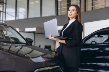 Bir arabanın yanında dizüstü bilgisayar tutan kadın, modern bir otomobil bayii ortamında müşterilerle meşgul oluyor.