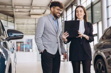 Bir galeride bir müşteriyle karşılaşan satıcı, elinde bir not panosu olan ve yeni bir arabanın özelliklerini tartışırken gülümseyen kadın.